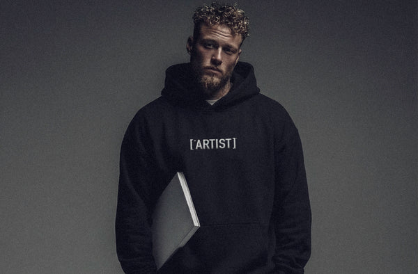 Model wearing ['ARTIST]™ hoodie in dark tone, minimal studio background
