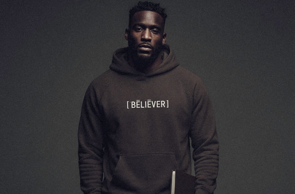 Model wearing ['BĒLIEVER]™ hoodie in dark tone, minimal studio background