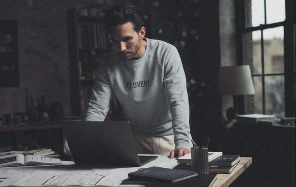 Man in a blue sweatshirt with 'LOVER' printed on it using a laptop at a desk.