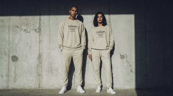 Two people wearing matching beige outfits with 'League' branding against a textured wall.